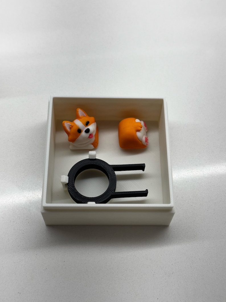 Op de afbeelding: Een witte doos bevat twee oranje en witte corgi-vormige keycaps en een zwarte keycap-trekker. E&eacute;n keycap toont het gezicht van de corgi en de andere zijn achterwerk. De keycap-trekker is zwart.