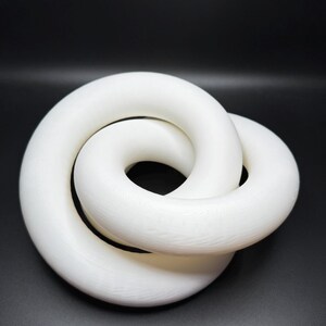 May include: A white, intertwined torus sculpture against a black background. The sculpture has a smooth, matte finish and a complex, knotted design. The form is a continuous loop, creating a visually interesting piece.