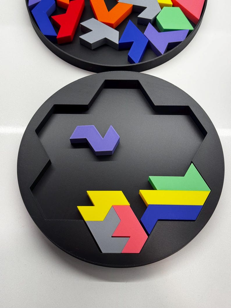 May include: A circular puzzle board with a hexagonal center. The board is black and has a variety of colorful geometric shapes that fit into the center. The shapes are purple, blue, yellow, green, gray, and pink.