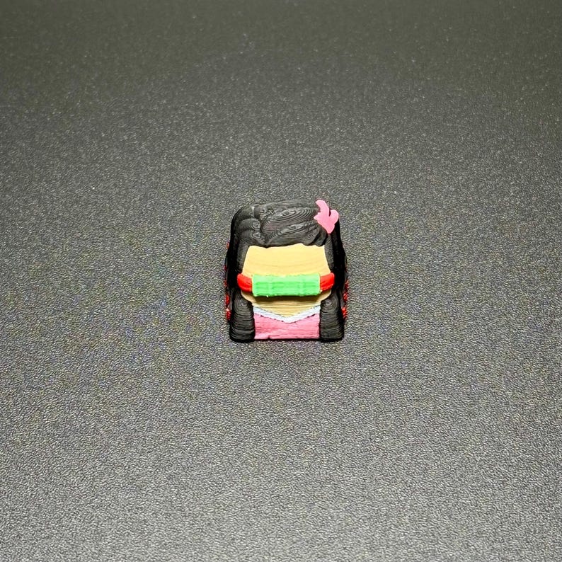 May include: A 3D printed keycap featuring a cartoon character with black hair, a pink bow, and a green mask. The character is wearing a pink dress.