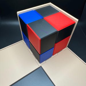 May include: A wooden box with a lid that opens to reveal a cube made of smaller cubes. The smaller cubes are red, blue, and black.
