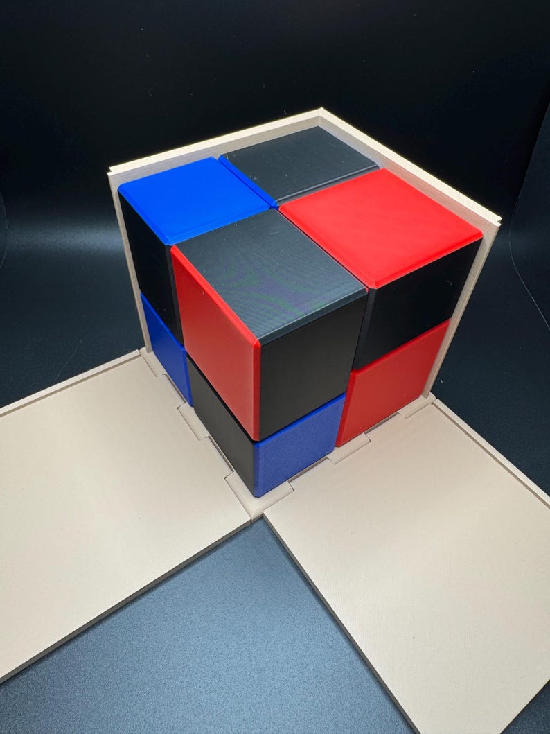 May include: A wooden box with a lid that opens to reveal a cube made of smaller cubes. The smaller cubes are red, blue, and black.