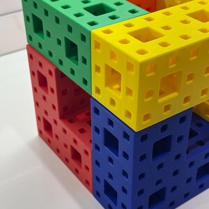May include: A multi-colored cube made of interlocking plastic pieces. The cube is made of four different colors: green, yellow, red, and blue.