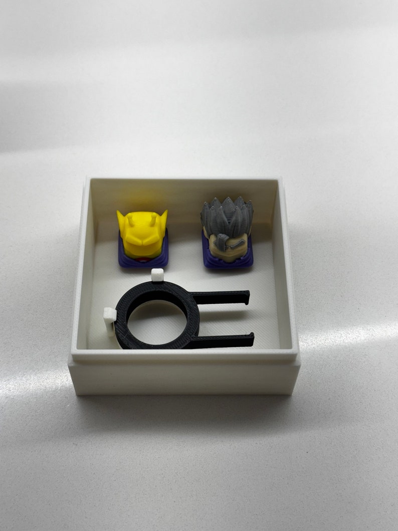 May include: A white box containing a black ring and two keycaps. One keycap is yellow and the other is grey.