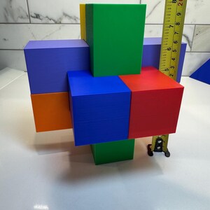 May include: A colorful wooden block puzzle with six blocks in different colors: blue, green, red, orange, yellow, and purple. The blocks are arranged in a cross shape, with the blue block in the center. The blocks are measured with a tape measure.
