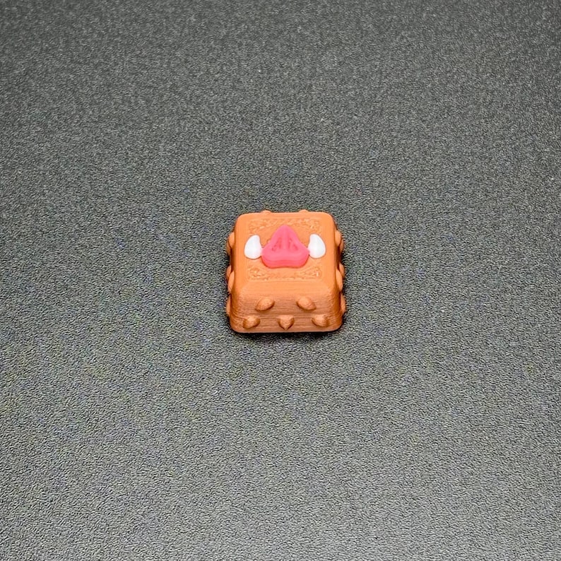 May include: A brown keycap with a white and pink pig face design. The keycap has a textured surface and is designed for mechanical keyboards.