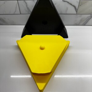 May include: A yellow triangular box with a lid and a small round knob on top. The box is sitting on top of a black triangular box.