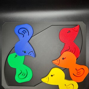 May include: A colorful wooden puzzle featuring six duck-shaped pieces in blue, green, red, orange, and yellow. The puzzle pieces are arranged on a black base with a curved design. The ducks have detailed features.