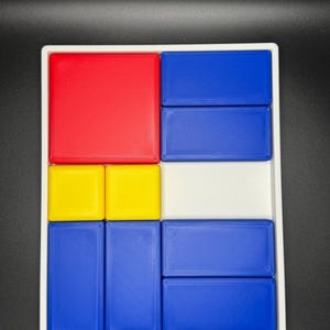 Op de afbeelding: Een wit dienblad met een geometrisch patroon in rood, geel en blauw. Het dienblad is verdeeld in negen secties, elk met een blok in een andere kleur.