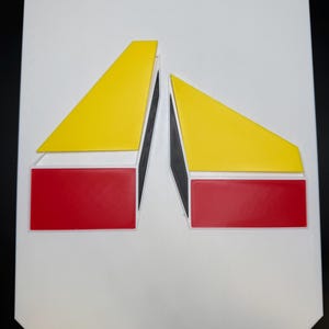 May include: A white rectangular sign featuring a geometric design. The design includes two yellow triangles and two red rectangles, all outlined in white and black. The shapes are arranged symmetrically against the white background.