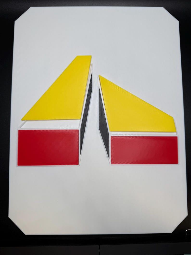 May include: A white rectangular sign featuring a geometric design. The design includes two yellow triangles and two red rectangles, all outlined in white and black. The shapes are arranged symmetrically against the white background.