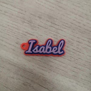 K&ouml;nnte beinhalten: Ein rotes und blaues Plastik-Namensschild mit dem Namen "Isabel" in lila.