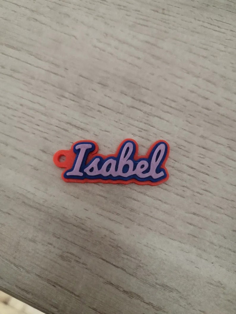 K&ouml;nnte beinhalten: Ein rotes und blaues Plastik-Namensschild mit dem Namen "Isabel" in lila.