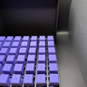 Może przedstawiać: Zestaw fioletowych stempli alfabetycznych zorganizowanych w czarnym plastikowym pudełku. Stemple są ułożone w kolejności alfabetycznej.