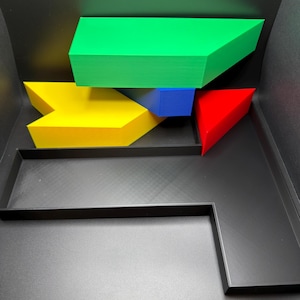 May include: A black plastic tray containing several geometric 3D printed shapes. The collection includes a large green trapezoidal block, a yellow arrow-shaped piece, a small blue rectangular prism, and a red triangular wedge arranged inside.