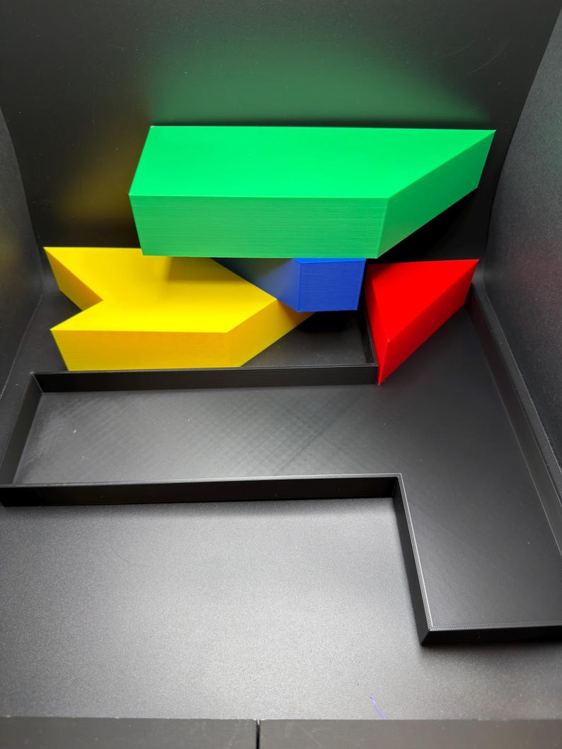 May include: A black plastic tray containing several geometric 3D printed shapes. The collection includes a large green trapezoidal block, a yellow arrow-shaped piece, a small blue rectangular prism, and a red triangular wedge arranged inside.