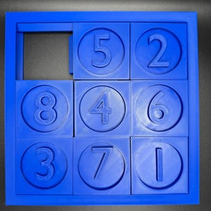 May include: A blue plastic puzzle with nine squares, each containing a circle with a number inside. The numbers are 5, 2, 8, 4, 6, 3, 7, and 1. One square is empty.