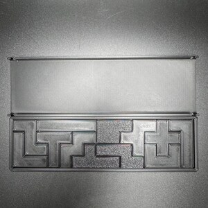 May include: A black plastic box with a sliding lid, containing a puzzle made of interlocking pieces. The puzzle is made of a gray plastic material and has a textured surface.