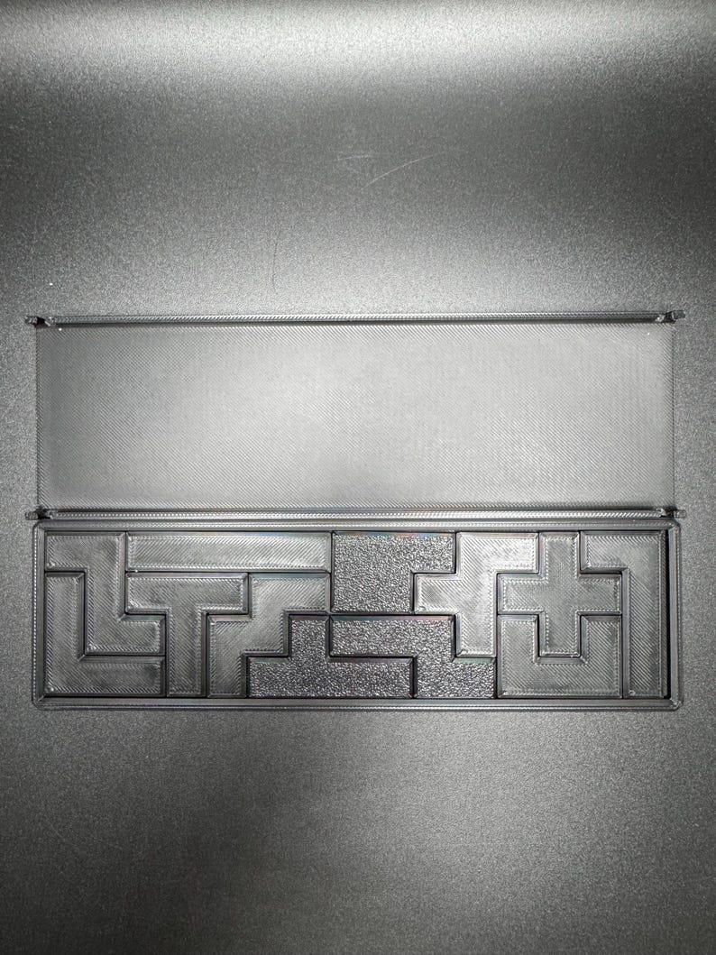 May include: A black plastic box with a sliding lid, containing a puzzle made of interlocking pieces. The puzzle is made of a gray plastic material and has a textured surface.