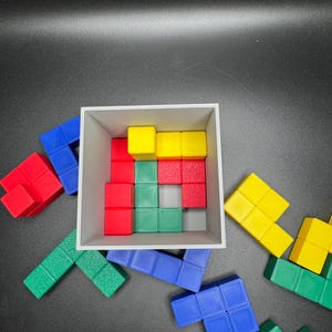 Puede incluir: Un conjunto de bloques de rompecabezas de pl&aacute;stico de colores en rojo, azul, verde y amarillo. Los bloques est&aacute;n dispuestos en forma de cuadrado, con algunos bloques dentro de una caja blanca y otros dispersos alrededor de la caja.