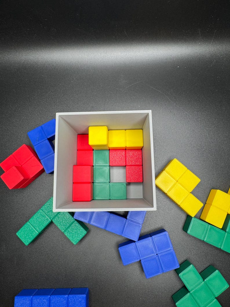 Puede incluir: Un conjunto de bloques de rompecabezas de pl&aacute;stico de colores en rojo, azul, verde y amarillo. Los bloques est&aacute;n dispuestos en forma de cuadrado, con algunos bloques dentro de una caja blanca y otros dispersos alrededor de la caja.