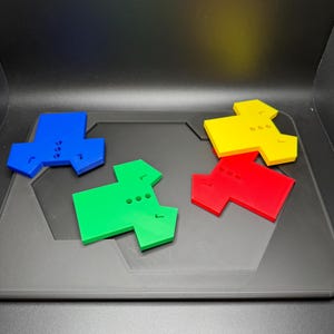 Puede incluir: Cuatro piezas de juego geom&eacute;tricas de colores azul, verde, rojo y amarillo. Cada pieza tiene una forma &uacute;nica con tres peque&ntilde;os agujeros y una peque&ntilde;a muesca. Las piezas est&aacute;n dispuestas sobre una superficie gris oscuro.