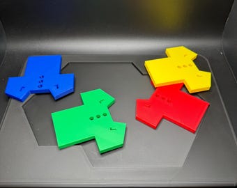Może przedstawiać: Cztery kolorowe, geometryczne elementy gry w kolorze niebieskim, zielonym, czerwonym i żółtym. Każdy element ma unikalny kształt z trzema małymi otworami i małym wcięciem. Elementy są ułożone na ciemnoszarej powierzchni.