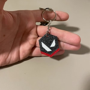 May include: A black and red hexagonal keychain with a silver key ring. The keychain features a design with white angular shapes on a black background, and a red border. The keychain is held in a hand.