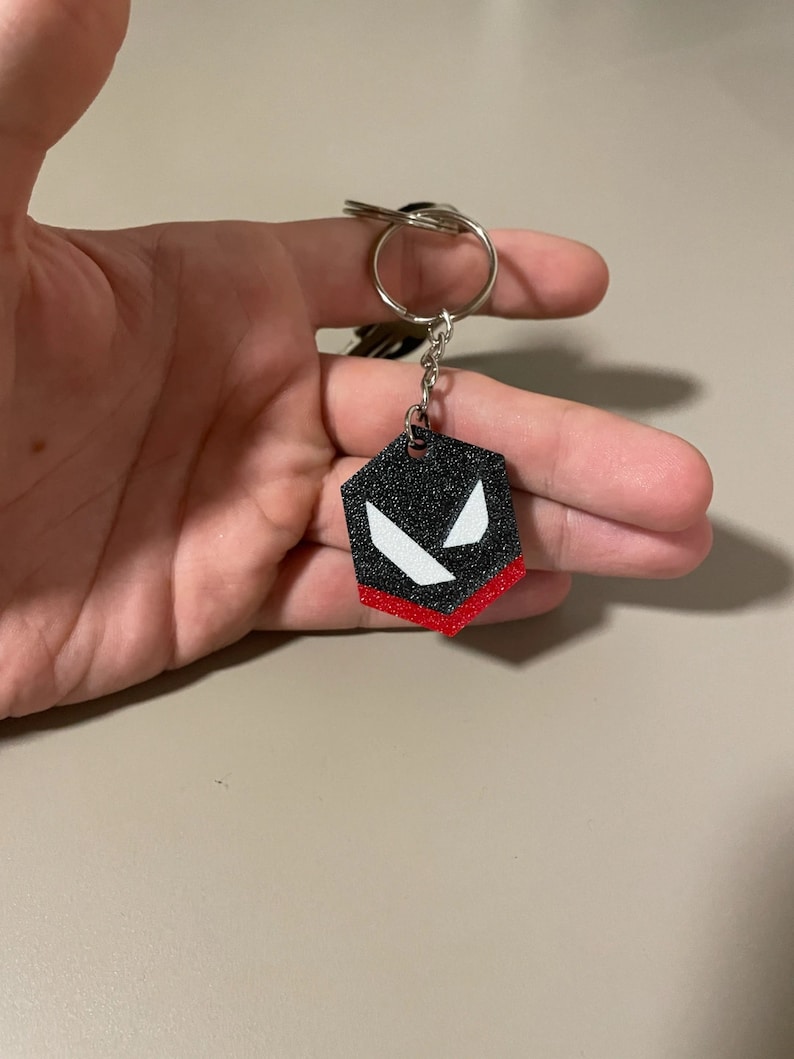 May include: A black and red hexagonal keychain with a silver key ring. The keychain features a design with white angular shapes on a black background, and a red border. The keychain is held in a hand.