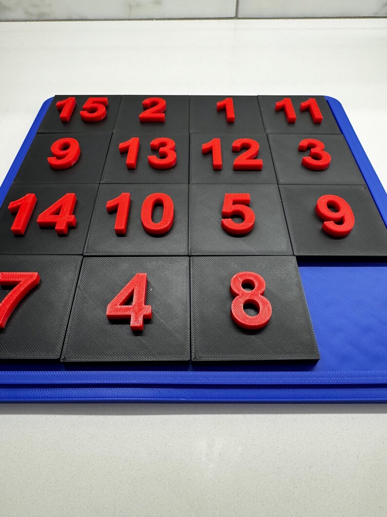 K&ouml;nnte beinhalten: Ein schwarzes und blaues Puzzlespiel mit roten Zahlen von 1 bis 15 auf jeder Kachel. Die Kacheln sind in einem 4x4-Raster angeordnet, wobei eine Kachel fehlt.