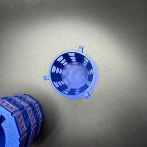 May include: A top-down view of a blue 3D-printed cylindrical container with a textured surface and three small handles. The container is next to a blue and purple geometric object.