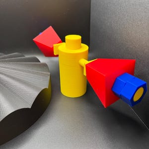 May include: A yellow cylindrical object with a flat top and two red and blue geometric shapes attached. A black, fan-shaped object is in the background. The objects are 3D printed.