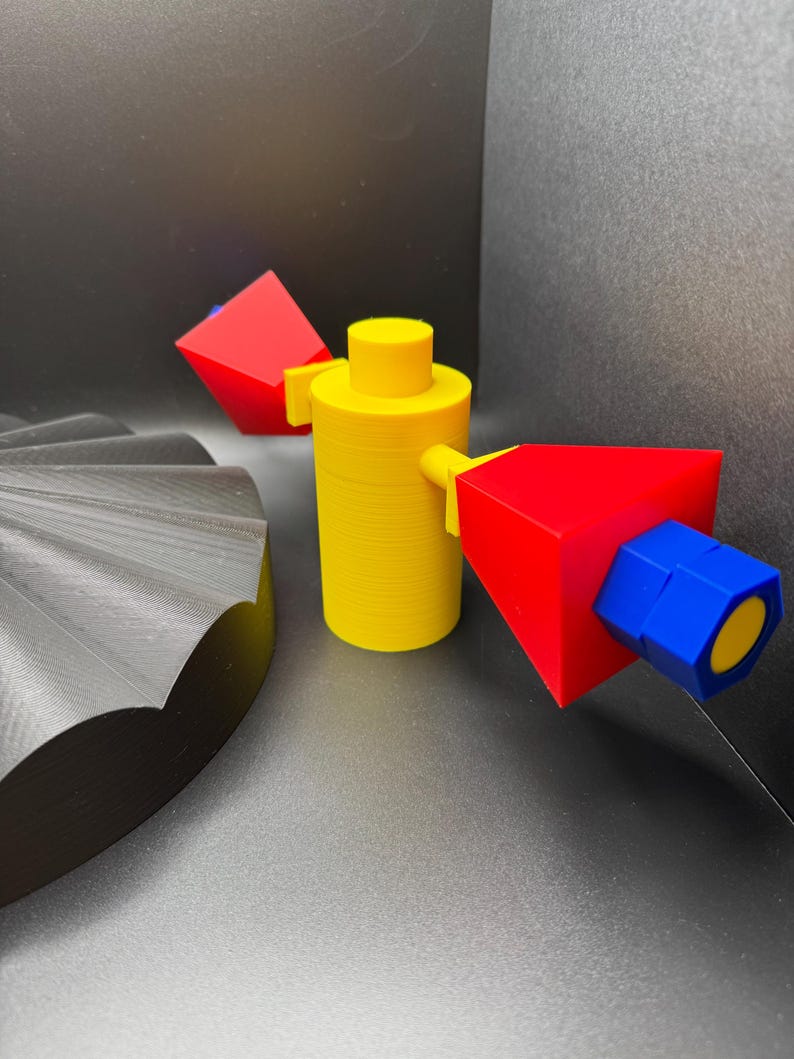 May include: A yellow cylindrical object with a flat top and two red and blue geometric shapes attached. A black, fan-shaped object is in the background. The objects are 3D printed.