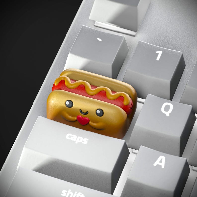May include: A gold hot dog keycap with a smiling face and a red heart, sitting on a white keyboard with the word "caps" printed on the key.