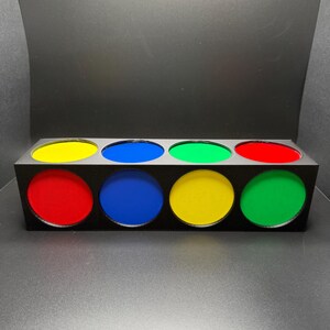 May include: A black rectangular box with eight circular apertures, each filled with a different colour: red, yellow, blue, and green. The box is on a grey surface with a black background.