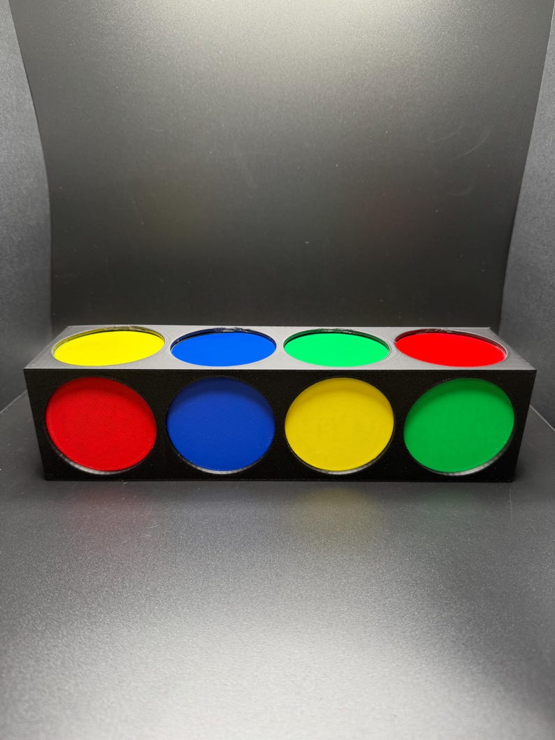 May include: A black rectangular box with eight circular apertures, each filled with a different colour: red, yellow, blue, and green. The box is on a grey surface with a black background.