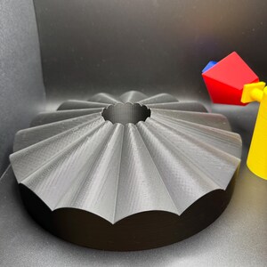 May include: A gray, 3D-printed bundt cake pan with a fluted design. The pan has a circular base and a central hole. A colorful toy is partially visible in the background.