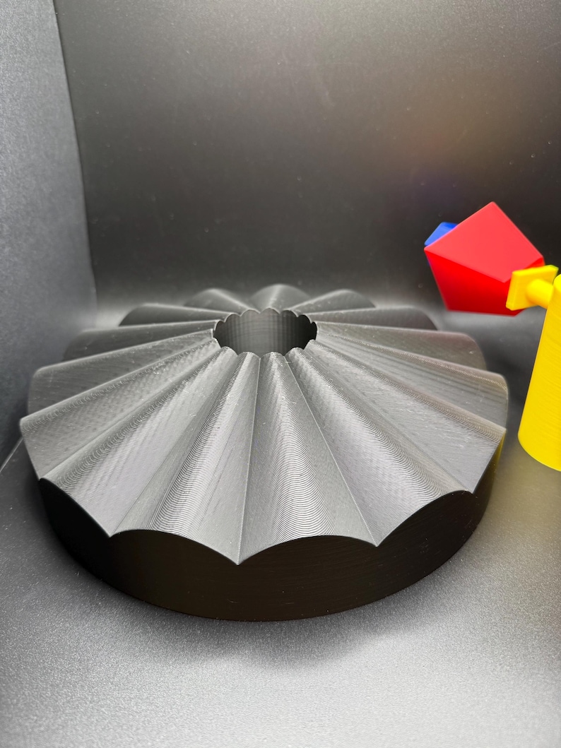 May include: A gray, 3D-printed bundt cake pan with a fluted design. The pan has a circular base and a central hole. A colorful toy is partially visible in the background.
