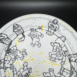 Puede incluir: Un plato blanco con una ilustraci&oacute;n en negro y amarillo de astronautas con trajes espaciales. Los astronautas est&aacute;n en varias poses, algunos de pie y otros flotando. El plato tiene un borde gris.