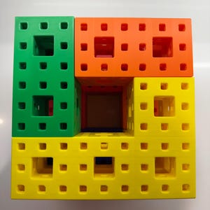 May include: A set of four colorful plastic building blocks, each with a grid of small holes. The blocks are stacked to form a square, with the top block being orange, the second block being green, the third block being yellow, and the bottom block being blue.