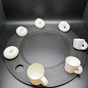 Op de afbeelding: Een verzameling kleine, witte keramische donuts en koffiemokken gerangschikt op een donkergrijze cirkelvormige tray. De donuts zijn ringvormig en de mokken hebben oren. De items zijn gelijkmatig verdeeld.