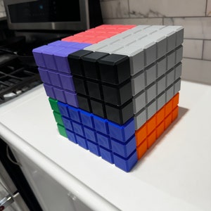 May include: A cube made of six different colored blocks. The blocks are arranged in a pattern, with the top block being gray, the front block being orange, the right side block being blue, the left side block being green, the back block being black, and the bottom block being purple.