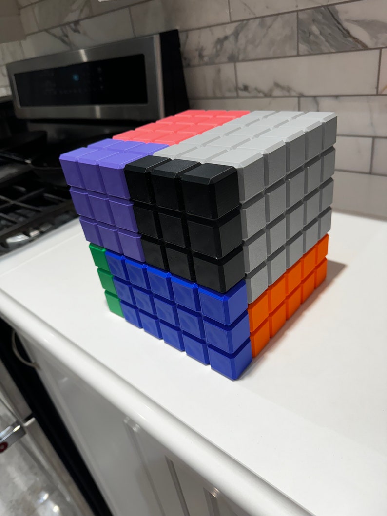 May include: A cube made of six different colored blocks. The blocks are arranged in a pattern, with the top block being gray, the front block being orange, the right side block being blue, the left side block being green, the back block being black, and the bottom block being purple.