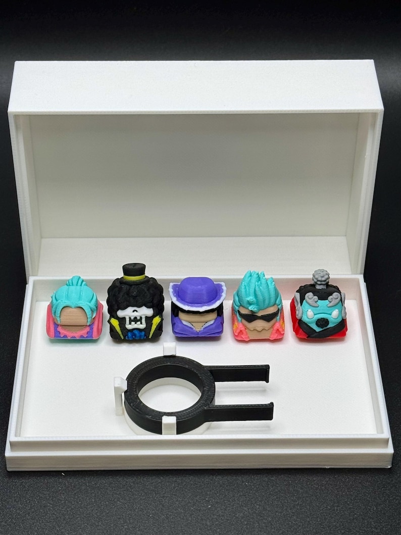 May include: A white box with a black ring and five colorful 3D printed keycaps featuring characters from the anime One Piece. The keycaps are Luffy, Brook, Sanji, Zoro, and Franky.