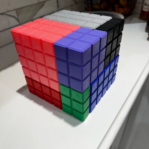 May include: A colorful cube puzzle made of small square blocks. The cube is made up of six different colors: red, purple, green, blue, black, and white.