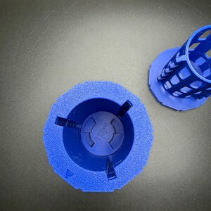 May include: Two blue, 3D-printed objects. One is a cylindrical cage with a hexagonal base. The other is a wider, hexagonal base with a central circular indentation. The word "TOP" is visible in the center of the base.