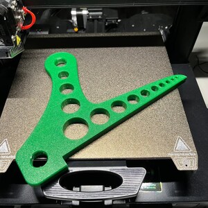 Może przedstawiać: Zielony plastikowy obiekt wydrukowany w 3D z wieloma otworami. Obiekt leży na teksturowanej powierzchni. Na powierzchni widoczny jest tekst "Warning hot surface".