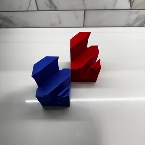 May include: Two 3D printed geometric shapes, one red and one blue, are stacked on a white surface. The shapes are made of plastic and have a smooth finish.