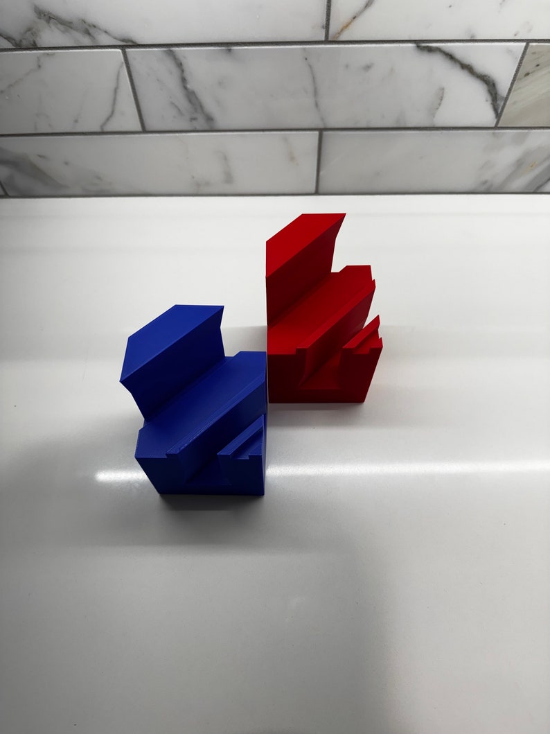 May include: Two 3D printed geometric shapes, one red and one blue, are stacked on a white surface. The shapes are made of plastic and have a smooth finish.
