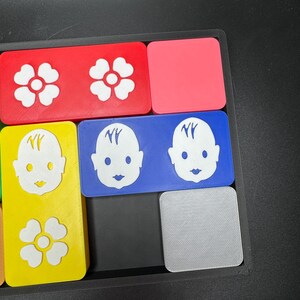 May include: A set of colorful square tiles with different designs. The tiles feature a white flower, a baby face with the letters "NY" on the forehead, and a gray tile with a textured surface.